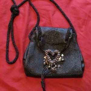 May include: A black leather purse with a beaded strap and a heart-shaped metal embellishment. The purse has a drawstring closure and a tassel.