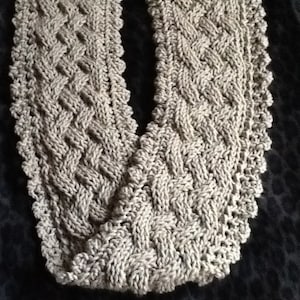 Puede incluir: Una bufanda de punto beige con un patrón trenzado y un borde festoneado.