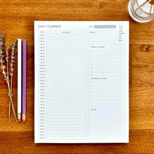May include: A white daily planner with a light blue grid layout, featuring sections for time, schedule, goals, and to-do lists. Two pencils and a sprig of lavender rest beside the planner on a wooden surface. A clear glass bottle is in the background.