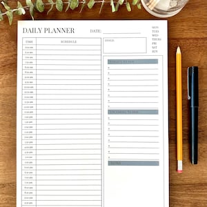 Puede incluir: Un planificador diario blanco con el texto "DAILY PLANNER" en la parte superior. El planificador tiene secciones para la hora, el horario, los objetivos y las listas de tareas. Un lápiz amarillo y un bolígrafo negro están junto al planificador sobre una superficie de madera.
