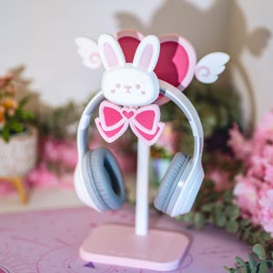 Può includere: Un supporto per cuffie rosa e bianco con un design a forma di coniglio. Il supporto ha una base a forma di cuore e un fiocco sulla testa del coniglio.