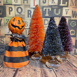 May include: A Halloween-themed display featuring a jack-o'-lantern figure and three bottle brush trees. The jack-o'-lantern has a smiling face and is atop a striped orange and black dress. The trees are orange, black, and purple, with glitter accents.