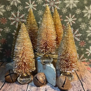 May include: A collection of gold-colored, glittery bottle brush Christmas trees of varying sizes, arranged with small, rustic bells and a clear glass ornament. The trees are set on a wooden surface with a snowflake patterned background.