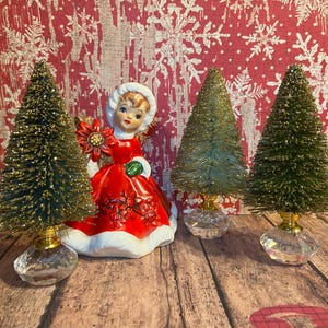 May include: A Christmas scene featuring a vintage figurine of a girl in a red dress holding a poinsettia, flanked by three miniature bottle brush Christmas trees. The trees have gold glitter and sit on crystal bases.