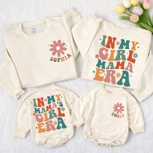 May include: Cream-colored matching family sweatshirts and rompers. The sweatshirts feature a retro flower graphic with the name "Sophia" and the phrase "In My Girl Mama Era." The rompers have the same design, with the name "Emily" on one.