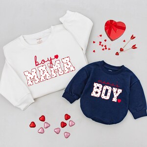 May include: Two sweatshirts, one white and one navy blue, with Valentine's Day themed text. The white sweatshirt says "boy MAMA" in red script and block letters. The navy blue sweatshirt says "mama's BOY". Scattered heart-shaped candies and a heart-shaped box.