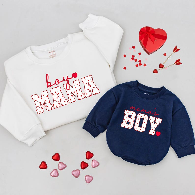 May include: Two sweatshirts, one white and one navy blue, with Valentine's Day themed text. The white sweatshirt says "boy MAMA" in red script and block letters. The navy blue sweatshirt says "mama's BOY". Scattered heart-shaped candies and a heart-shaped box.