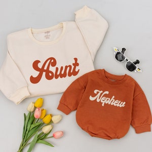 May include: A set of two cream colored sweatshirts with the words "Aunt" and "Nephew" in a retro font. The "Aunt" sweatshirt is on a white background and the "Nephew" sweatshirt is on a rust colored background.