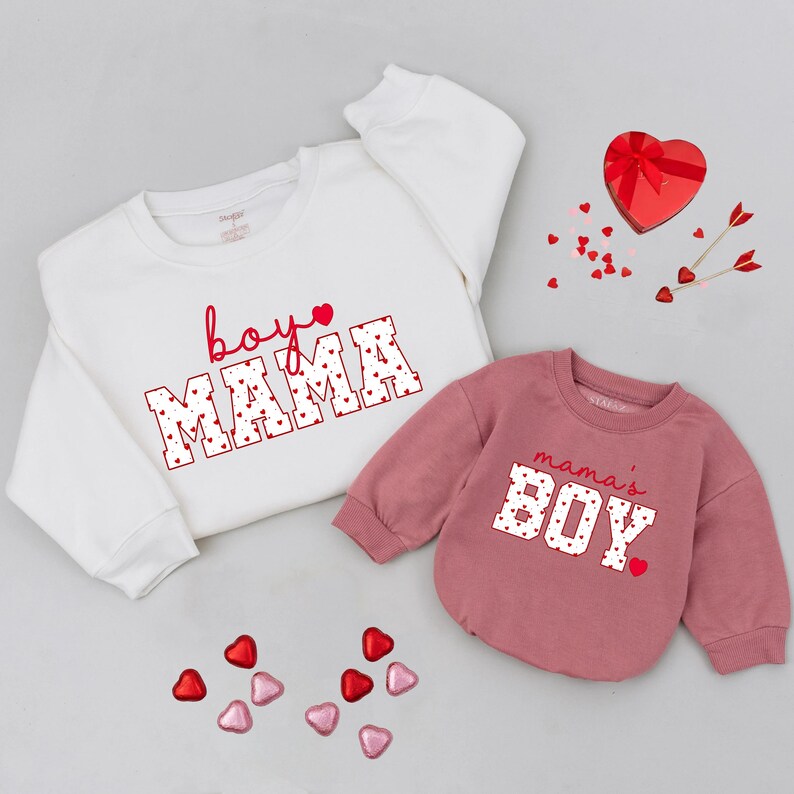 May include: Two sweatshirts, one white and one dusty rose, with Valentine's Day themed text. The white sweatshirt says "boy mama" in red lettering with heart accents. The dusty rose sweatshirt says "mama's boy" in white lettering with red heart accents.