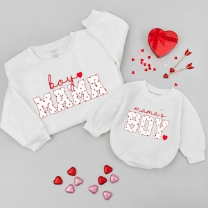 May include: Two white sweatshirts, one adult and one infant, with red text and heart accents. The adult sweatshirt says "boy MAMA" and the infant one says "mama's BOY". Scattered heart-shaped candies and a heart-shaped box are also present.