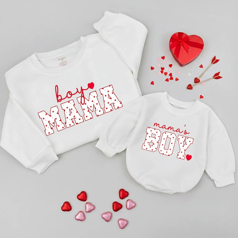May include: Two white sweatshirts, one adult and one infant, with red text and heart accents. The adult sweatshirt says "boy MAMA" and the infant one says "mama's BOY". Scattered heart-shaped candies and a heart-shaped box are also present.