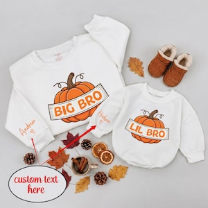 May include: Two white long-sleeve sweatshirts featuring an orange pumpkin graphic with a banner reading BIG BRO and LIL BRO. The sleeves are personalized with the names Andrew and Hudson in orange script, each accompanied by a small heart icon.