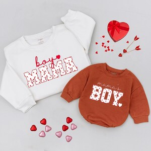 May include: Two sweatshirts, one white and one rust-colored, with Valentine's Day themed text. The white sweatshirt says "boy mama" in red script and block letters filled with hearts. The rust-colored one says "mama's boy" in white block letters with heart details.