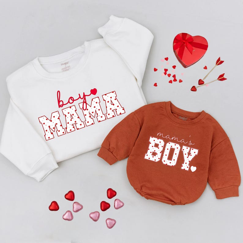 May include: Two sweatshirts, one white and one rust-colored, with Valentine's Day themed text. The white sweatshirt says "boy mama" in red script and block letters filled with hearts. The rust-colored one says "mama's boy" in white block letters with heart details.