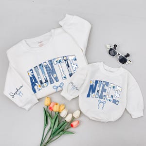 May include: White sweatshirts with blue floral and gingham lettering that spells "AUNTIE" and "NIECE". The "AUNTIE" sweatshirt has "Sophia" on the sleeve. Sunglasses and flowers are also in the image.