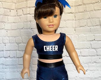 May include: A doll in a navy blue cheerleading outfit. The outfit features a crop top and shorts with black trim, and the word "CHEER" in white. The doll has brown hair and a blue bow.