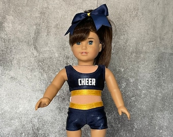 May include: A doll wearing a navy blue and gold cheerleading outfit with the word "CHEER" on the top. The doll has brown hair and blue eyes and is wearing a blue bow.
