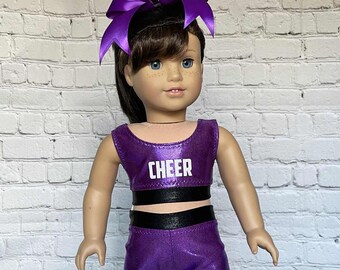 May include: A doll in a purple cheerleader outfit with the word "CHEER" in white on the top. The outfit includes a matching bow and shorts. The doll has brown hair and blue eyes.