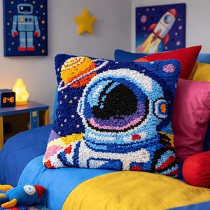 Può includere: Un cuscino quadrato con un design testurizzato a punch needle raffigurante un astronauta in tuta blu e bianca, su uno sfondo spaziale blu scuro con un pianeta giallo e arancione. Il design presenta colori vivaci e dettagli a rilievo.