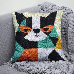 Könnte beinhalten: Ein farbenfrohes geometrisches Katzendesign auf einem Plüschkissen. Das Kissen besteht aus einem weichen, strukturierten Stoff und hat eine Farbgebung in Schwarz, Weiß, Orange, Grün und Gelb.