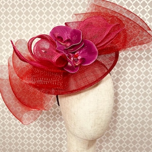 Op de afbeelding: Een felrode fascinator met een brede, gestructureerde rand van verstevigde gaasstof. Het hoofddeksel is versierd met een groep diepmagenta orchideeën die uit het midden zijn geplaatst, wat zorgt voor een gedurfd en gelaagd sculpturaal effect.