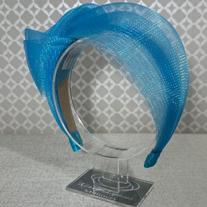 Puede incluir: Una diadema de color azul brillante con una tela de malla ancha, estructurada y semitransparente, que presenta un gran lazo superpuesto en un lado. La diadema se exhibe en un soporte de acrílico transparente con la etiqueta Kerry Lyons Millinery.