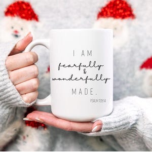 Może przedstawiać: Biały kubek ceramiczny z czarnym tekstem, który brzmi "I am fearfully & wonderfully MADE. PSALM 139:14". Kubek jest trzymany przez osobę ubraną w szary sweter z czerwonymi i białymi wzorami świątecznymi.