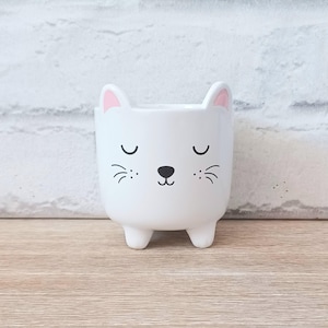 May include: A white ceramic cat-shaped planter with a black nose, whiskers, and closed eyes. The ears have pink inner details. The planter has three small feet and is set against a white brick wall and a wooden surface.