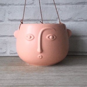 Può includere: Un vaso da fiori sospeso in ceramica rosa a forma di viso con un semplice naso, occhi e bocca. Il vaso ha un gancio in corda.