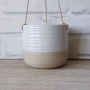 Puede incluir: Maceta colgante de cerámica blanca con fondo beige. La maceta tiene un diseño acanalado y está suspendida por una cuerda.