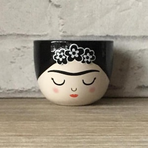 以下が含まれることがあります： 黒と白の小さなセラミック植木鉢。顔が描かれており、黒い上部、白い顔、閉じられた目、赤い口が特徴です。白い花の冠が植木鉢の上部を飾っています。