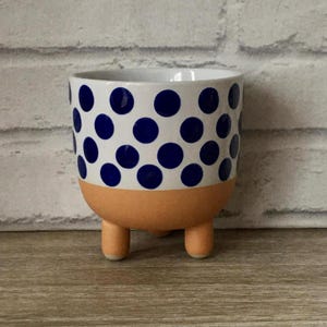 Puede incluir: Pequeña maceta de cerámica con base blanca y lunares azul marino. La sección inferior es naranja y tiene tres patas pequeñas y redondeadas. Adecuada para plantas pequeñas o suculentas, diseñada para uso en interiores.