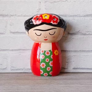 Könnte beinhalten: Eine Keramikfigur von Frida Kahlo in einem roten Kleid mit grünem Blumendruck und einer Blumenkrone. Die Figur hat einen verspielten Stil mit geschlossenen Augen und einem einfachen Lächeln.