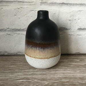Könnte beinhalten: Eine kleine, schwarze Keramikvase mit einer weißen, braunen und beigefarbenen Glasur. Die Vase hat einen runden Körper und einen schmalen Hals.
