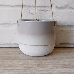 Puede incluir: Un macetero colgante de cerámica blanco y gris con un fondo texturizado y un efecto degradado rosa claro. El macetero tiene un colgador de cuerda.