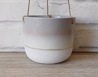 May include: A white and gray ceramic hanging planter with a textured bottom and a light pink ombre effect. The planter has a rope hanger.