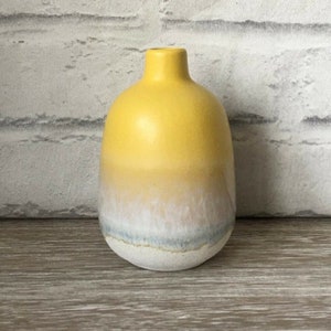 Może przedstawiać: Mały, żółty ceramiczny wazon o zaokrąglonym korpusie i wąskiej szyi. Wazon ma gradientowy wzór, przechodzący od żółtego u góry do białego i niebieskiego u podstawy. Wazon ma około 15 cm wysokości.
