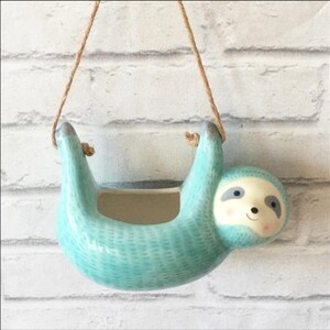 Puede incluir: Un macetero colgante de cerámica con forma de perezoso. El macetero es de color azul claro con una superficie texturizada y una cara de perezoso sonriente. Cuelga de una cuerda marrón. Ideal para plantas pequeñas.
