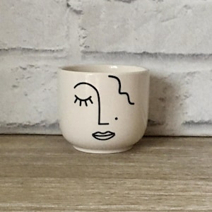 May include: A small, off-white ceramic cup with a minimalist face design in black. The design includes a closed eye, a nose, a mouth, and a wavy line. The cup is cylindrical and sits on a wooden surface.
