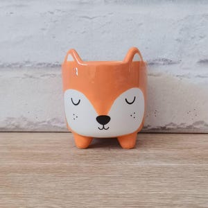 May include: A ceramic fox-shaped plant pot in orange, white, and black. The pot has a glossy finish, with the fox's face featuring closed eyes, a black nose, and small dots. The pot is designed with four small feet and two ears.