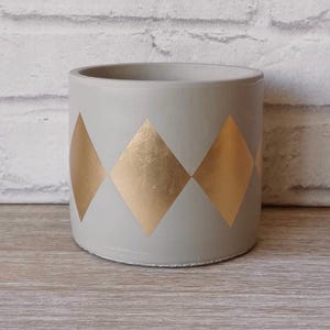 May include: A gray concrete planter with a geometric pattern of gold diamonds. The planter is a cylindrical shape and has a smooth, matte finish.