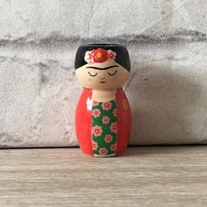 Puede incluir: Figura decorativa pintada a mano que representa a Frida Kahlo. La figura presenta un corte de pelo negro, un tocado floral y un vestido rojo con un corpiño verde con estampado floral. La figura está sobre una superficie de madera.