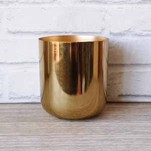 May include: A cylindrical, gold-colored metal container with a rounded base. The container has a smooth, reflective surface and is set against a white brick wall and a wooden surface. The container is likely a decorative item or a holder.