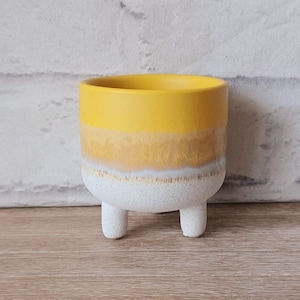 Peut inclure: Un pot en céramique jaune, blanc et marron avec trois pieds. Le pot est fait d'un matériau texturé et a une forme arrondie.