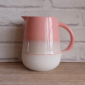Könnte beinhalten: Zweifarbiger Keramikkrug mit rundem Boden und Henkel. Die obere Hälfte ist glänzend rosa, geht unten in ein gesprenkeltes Weiß über. Der Krug hat einen Ausguss und steht auf einer Holzoberfläche.