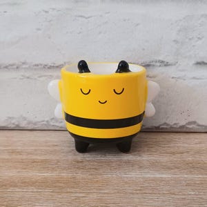 Puede incluir: Una maceta de cerámica amarilla con forma de abeja. La maceta tiene rayas negras, pequeñas patas negras y alas blancas. La cara de la abeja tiene los ojos cerrados y una boca sonriente. La maceta está sobre una superficie de madera.