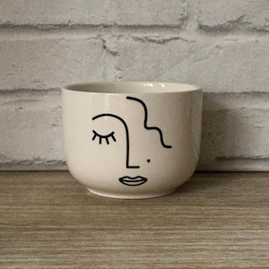 May include: A cream ceramic plant pot with a minimalist face design. The face is outlined in black, featuring a closed eye, a nose, a small dot, and a mouth. The pot has a rounded shape and sits on a wooden surface.