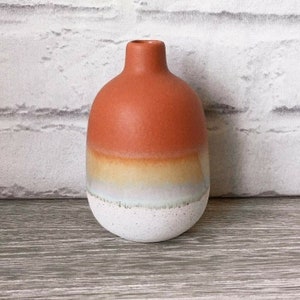 Könnte beinhalten: Eine kleine Keramikvase mit rundem Körper und schmalem Hals. Die Vase ist hauptsächlich orange, geht in Gelb, Weiß und gesprenkeltes Weiß am Boden über. Die Vase steht auf einer Holzoberfläche.