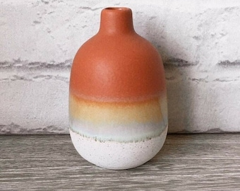 May include: A small, ceramic vase with a rounded body and a narrow neck. The vase is primarily orange, transitioning to yellow, white, and speckled white at the base. The vase is displayed on a wooden surface.