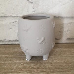 May include: A grey ceramic planter with a textured surface and a design of bees, flowers, and crowns. The planter has three small feet.